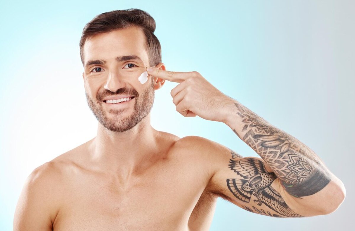 A muscular man with a detailed tattoo on his arm poses against a soft blue background, pointing to his temple thoughtfully.
