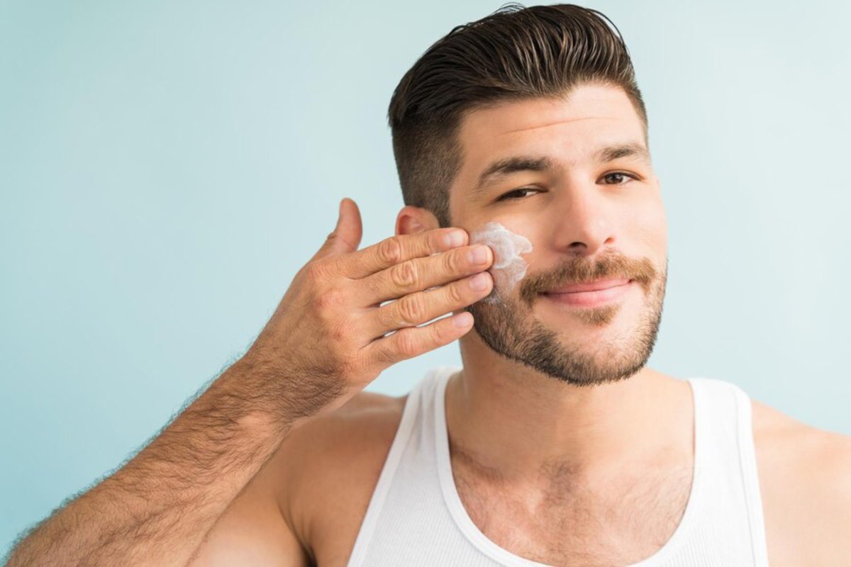 A man wearing a white tank top touches his face with one hand, set against a soft blue background.