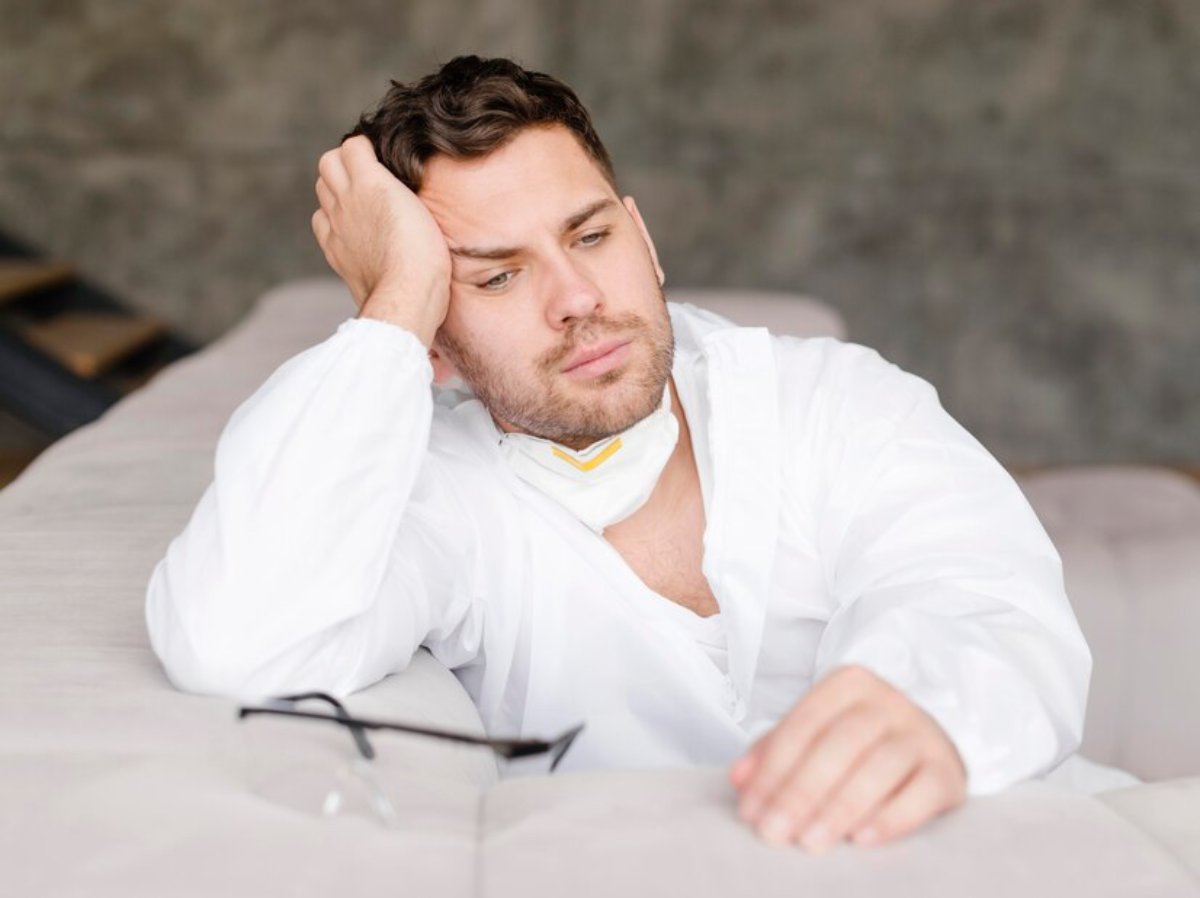 A person in protective clothing rests on a sofa, with one hand on their forehead and glasses lying nearby. The background features textured walls.