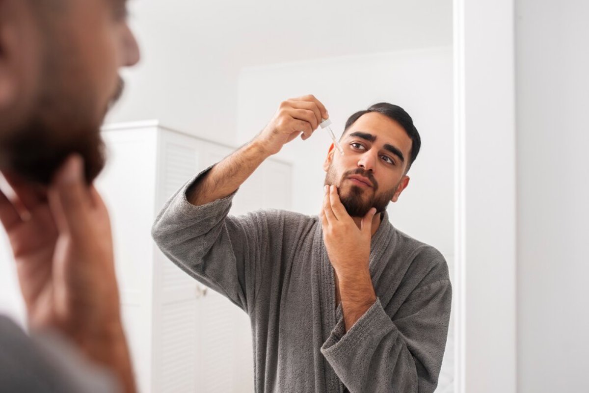 A person in a gray robe inspects their beard in a bright bathroom mirror, focusing on grooming and self-care.