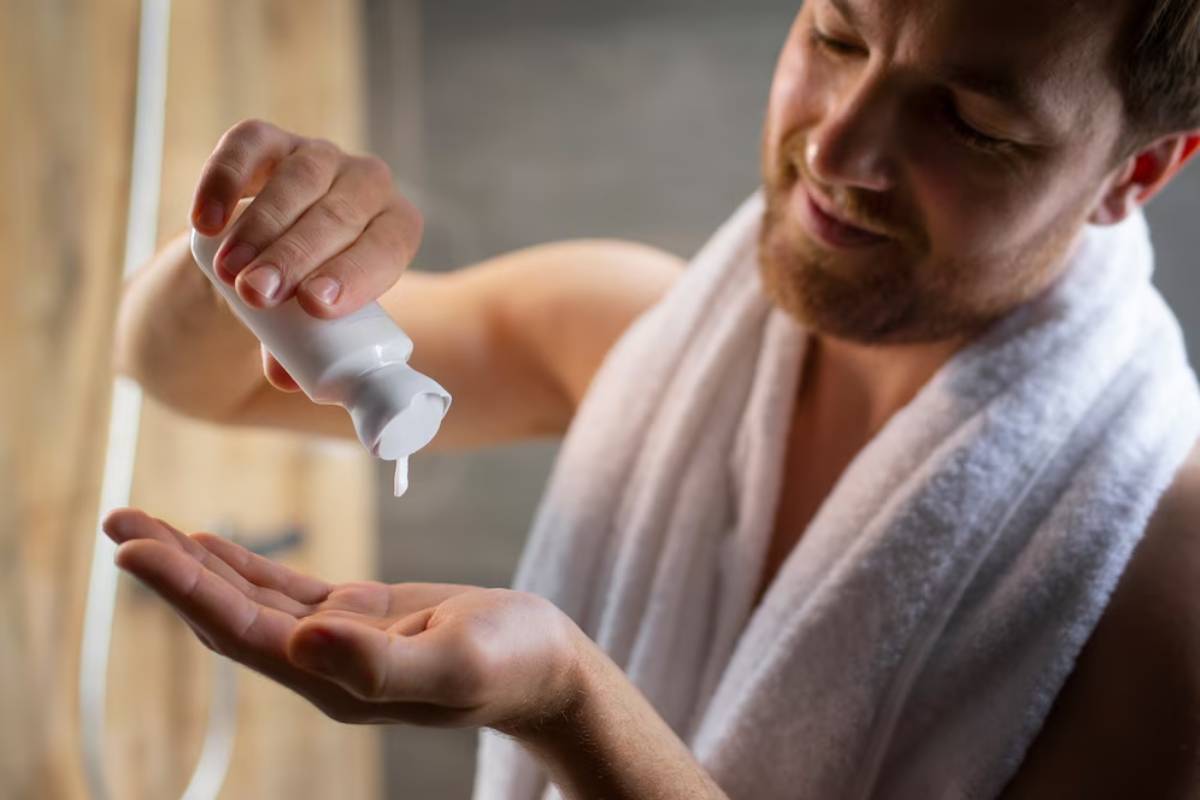 A person in a towel dispenses lotion from a small white bottle into their palm, in a modern, warmly lit bathroom environment.
