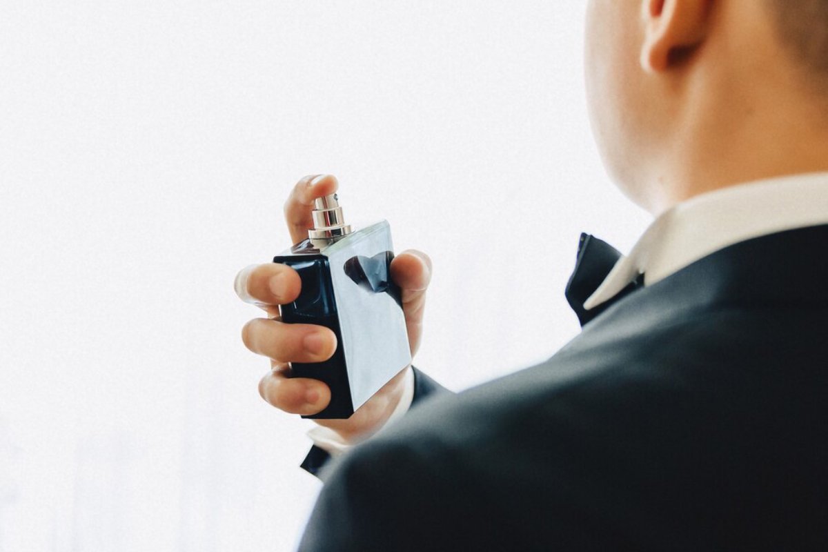 A man in a tuxedo holds a sleek, dark perfume bottle, preparing to apply it against a bright, light-filled background.