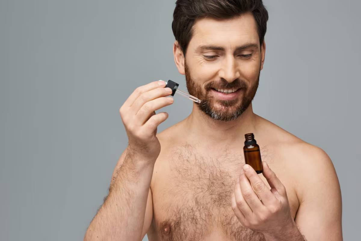 A bare-chested man holds an amber bottle and dropper, preparing to apply a serum or oil, against a neutral gray background.
