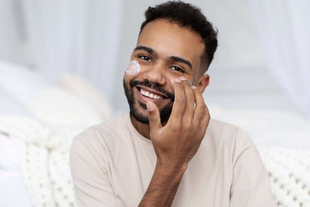 A person draped in a light-colored shirt is resting their chin on their hand, seated comfortably on a bed with soft, white bedding.