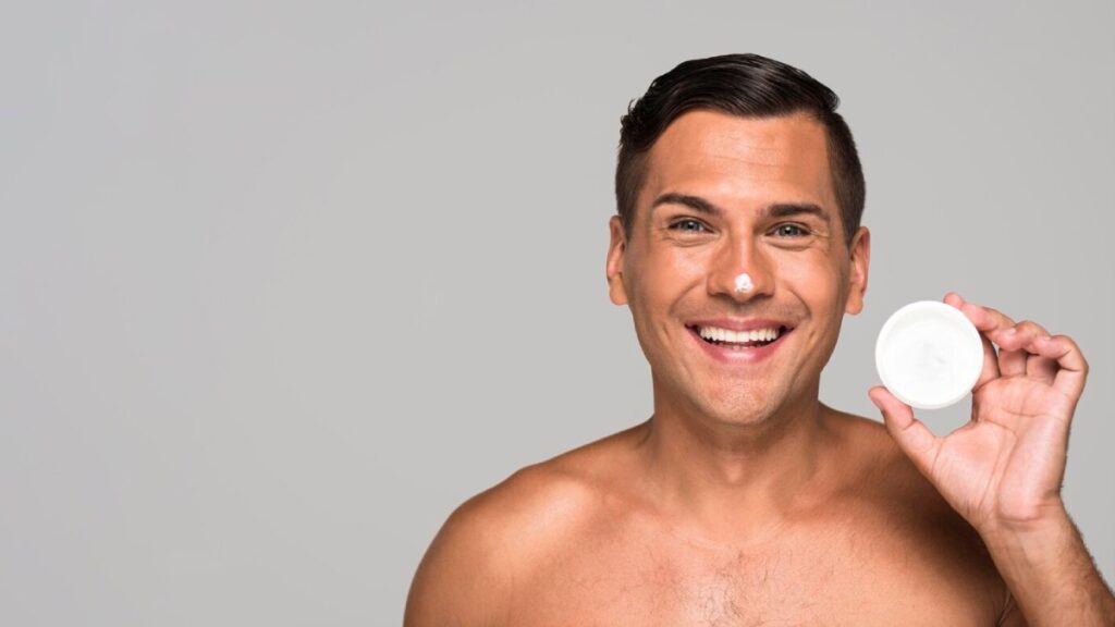 A muscular man holding a small white container in front of his chest, against a plain gray background.
