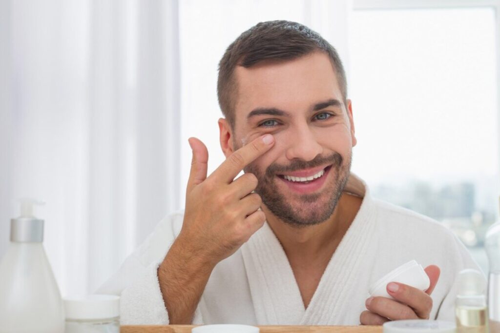 A person in a white bathrobe applies cream to their face in a bright, airy bathroom surrounded by skincare products.
