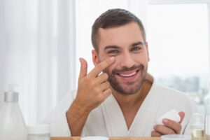 A person in a white bathrobe applies cream to their face in a bright, airy bathroom surrounded by skincare products.