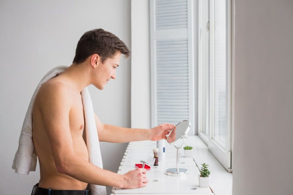 A shirtless man with a towel draped over his shoulder examines himself in a mirror at a well-lit vanity with small plants.