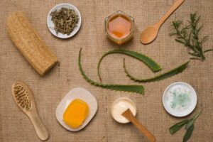 An assortment of natural beauty products: aloe vera leaves, herbal tea, honey, soap, lotion, and a wooden brush over a burlap background.