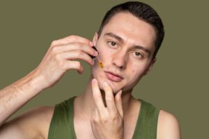 A person with short dark hair holds their hands near their face, wearing a green tank top against a muted olive background.
