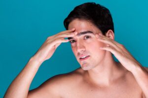 A young man with dark hair poses against a vibrant blue background, holding his hands near his head in a thoughtful gesture.