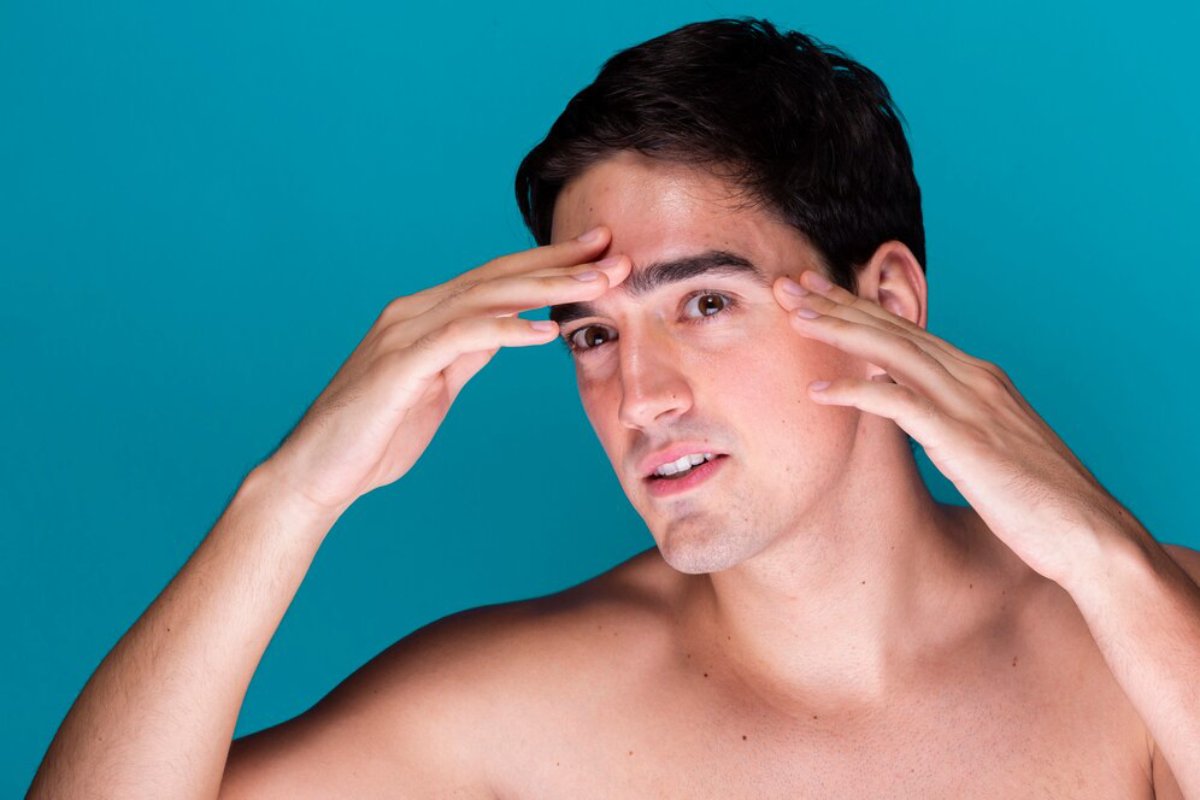 A young man with dark hair poses against a vibrant blue background, holding his hands near his head in a thoughtful gesture.