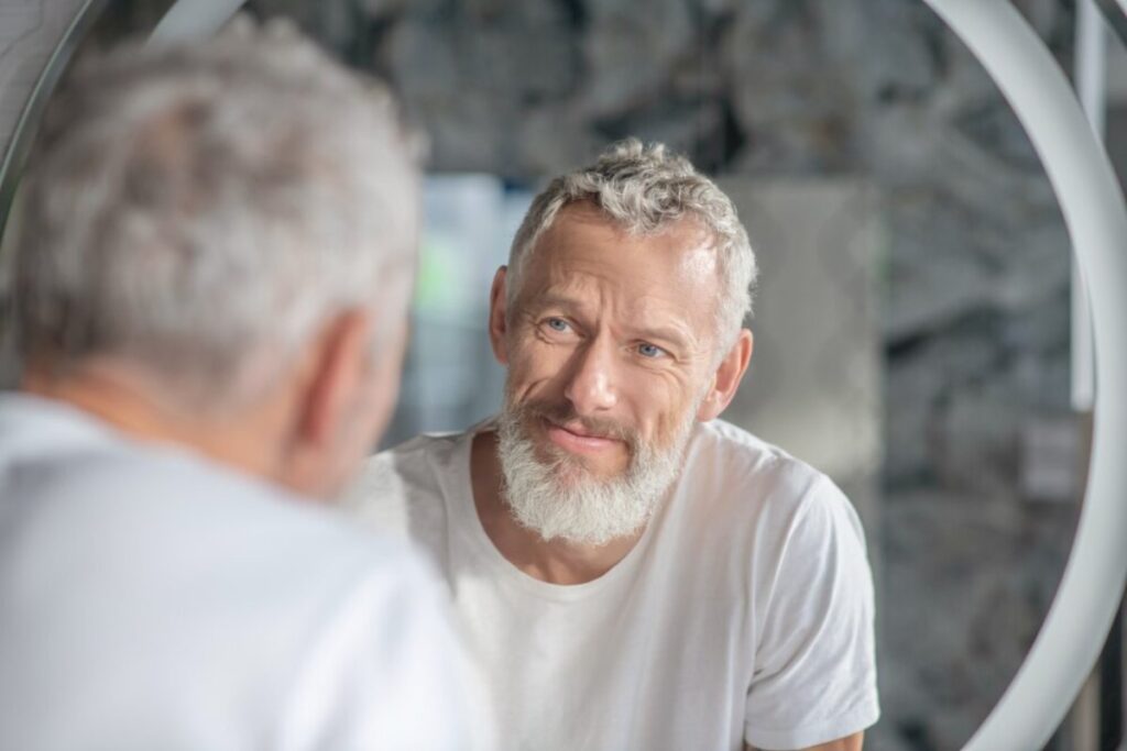 A man with grey hair and a beard sits in front of a circular mirror, reflecting a serene indoor space with stone walls.