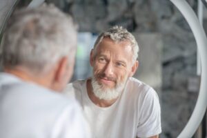 A man with grey hair and a beard sits in front of a circular mirror, reflecting a serene indoor space with stone walls.