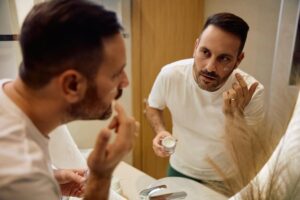 A man applies skincare cream in a stylish bathroom, gazing in the mirror while holding a small jar of cream.