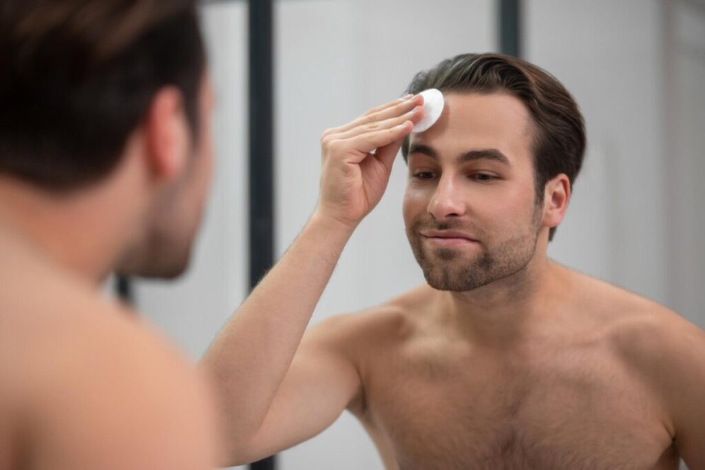 A shirtless man stands in front of a mirror, applying skincare product to his forehead. The background features a modern bathroom design.
