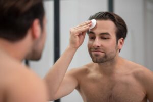 A shirtless man stands in front of a mirror, applying skincare product to his forehead. The background features a modern bathroom design.