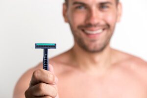 A person holds a blue razor with a green blade, positioned in front of a plain white background. The individual has an exposed torso.