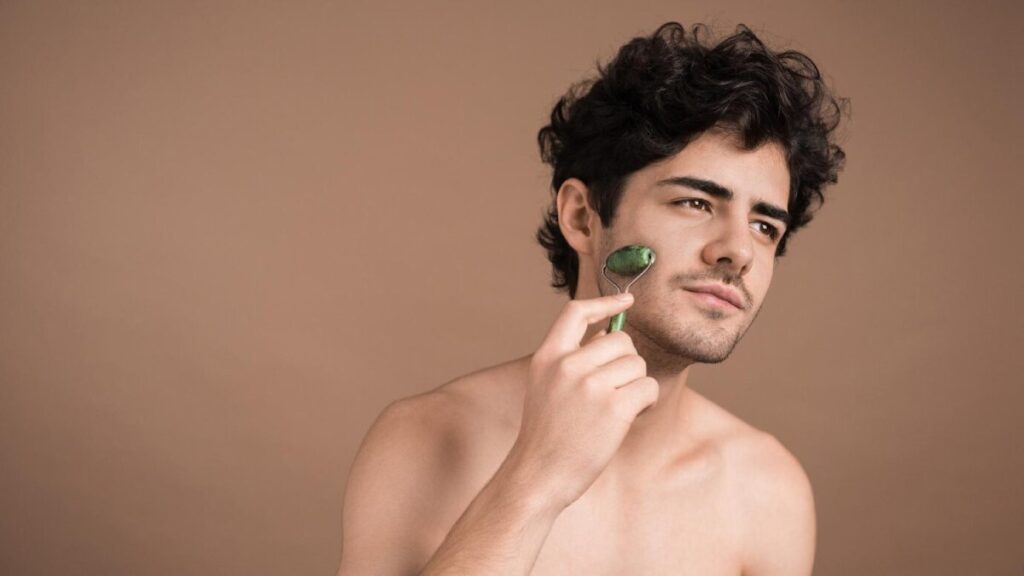 A young person with curly hair poses against a soft, neutral background, gently touching their chin with their fingers.