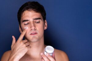 A shirtless individual holds a skincare jar in one hand while applying cream with the other, set against a blue backdrop.