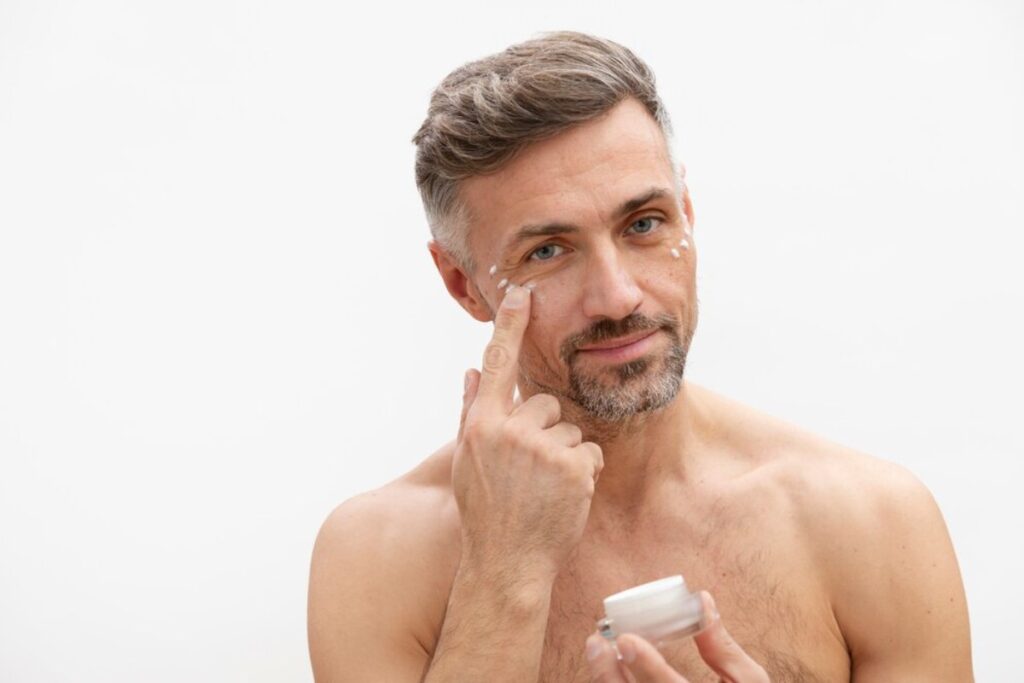 A shirtless man inspects a skincare cream jar, thoughtfully touching his chin against a minimalist white background.