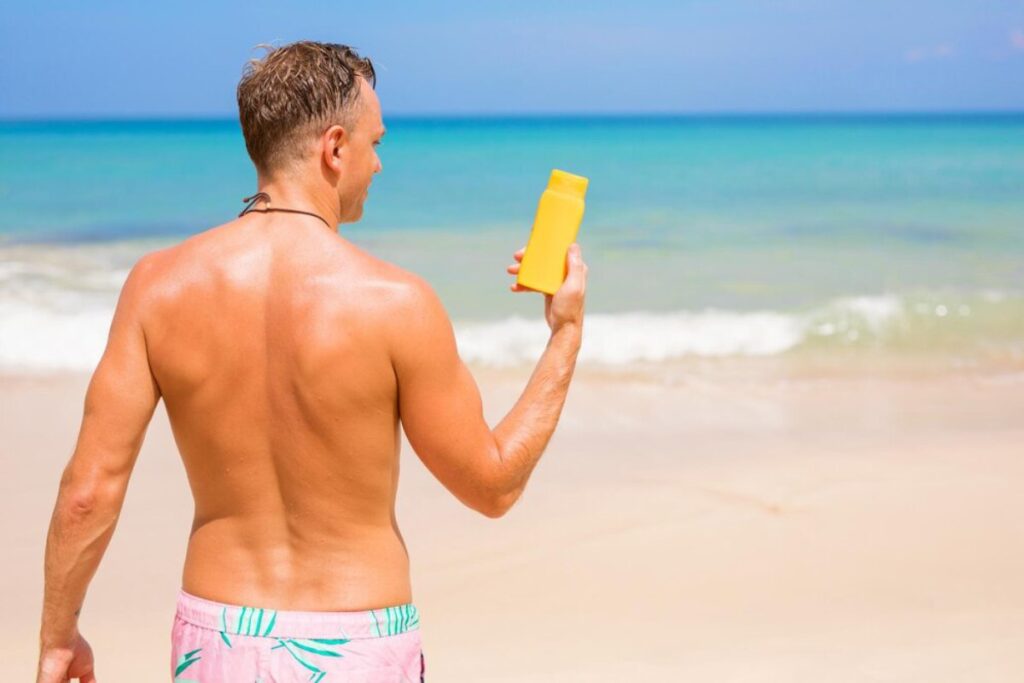 A man with a toned back stands on a sandy beach, holding a yellow sunscreen bottle, with ocean waves gently lapping behind him.