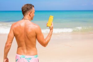 A man with a toned back stands on a sandy beach, holding a yellow sunscreen bottle, with ocean waves gently lapping behind him.