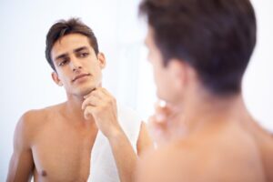A young man stands in front of a mirror, thoughtfully touching his chin while wearing a towel around his neck in a bright bathroom.
