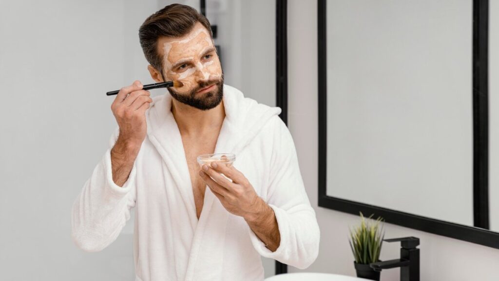 A man in a white bathrobe applies makeup using a brush, holding a small container in his other hand, with a modern bathroom setting.