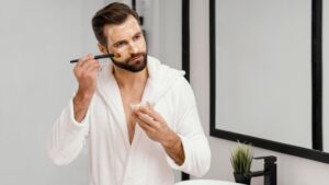 A man in a white bathrobe applies makeup using a brush, holding a small container in his other hand, with a modern bathroom setting.