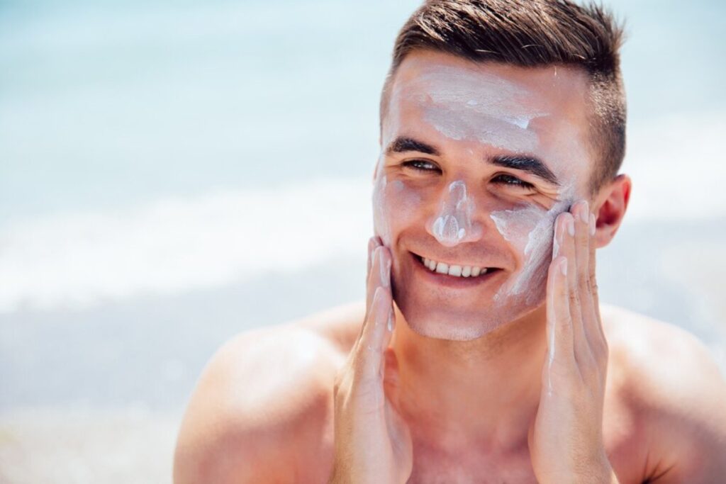 A young man at the beach, sitting on the sand, with the ocean in the background; he's holding his hands to his face.