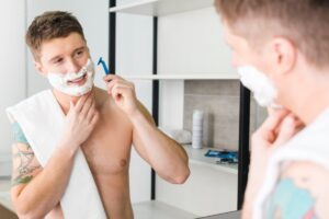 A shirtless young man with a towel around his neck holds a blue razor, gazing thoughtfully into a bathroom mirror.