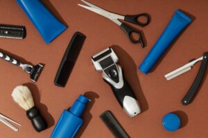A flat lay of grooming tools: razors, scissors, combs, brushes, and grooming products arranged on a brown background.