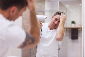 A person in a white shirt uses a hairdryer while looking into a bathroom mirror, with modern decor in the background.