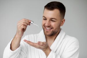 A person in a white robe holds a dropper bottle over their palm, preparing to apply a serum or skincare product.