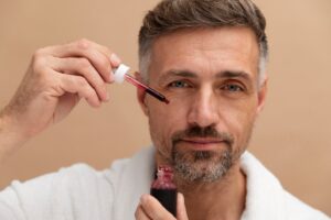 A person in a white robe applies a serum with a dropper to their skin, set against a soft beige background.